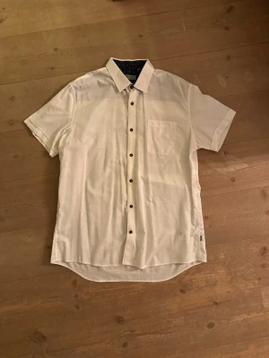 Vit kortärmad skjorta från Matinique - Säljer en stilren vit skjorta från Matinique med korta ärmar och bröstficka. Skjortan har mörka knappar och insidan av kragen är fodrad med marinblått tyg med små vita blommor. Passformen är slim och materialet känns lätt och skönt.