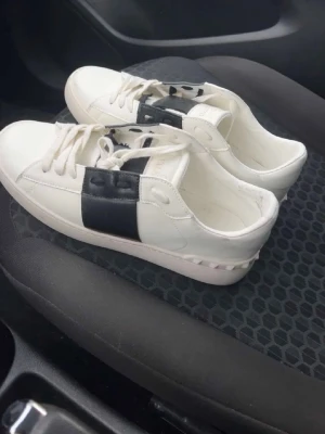 Valentino Open sneakers vit/svart - Säljer ett par Valentino Open sneakers i vitt skinn med bred svart läderdetalj över snörningen. Klassisk rund tå, platt sula och nitar bak på hälen för extra edge. Snygg och stilren modell som lyfter vilken outfit som helst.