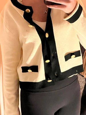 Beige och svart kofta med guldknappar - Snygg beige kofta med svarta detaljer och guldknappar framtill. Koftan har lång ärm, ribbade muddar och två dekorativa fickor med svarta band och guld knappar. Perfekt för dig som gillar klassisk och trendig stil. Jag använder den inte så därför väljer jag att sälja den. Tyvärr har den dock fått en liten svart prick där fram ( se bild 4). Själv är jag i storlek xs men denna kofta passar mig ändå, även fast den är en storlek större.❣️