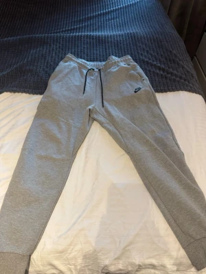 Nike Tech Fleece Grå storlek M - Säljer ett par grå mjukisbyxor från Nike i storlek M. Byxorna har resår i midjan med svart snörning, ribbade muddar vid bensluten och en diskret Nike-logga på vänster lår. Perfekta för chill eller träning. De är nytvättade. 