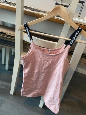 Rosa linnetopp med svarta band XS - Säljer en ljusrosa linnetopp med svarta axelband där det står 'CALIFORNIA' i vitt. Toppen har enkel, rak passform och ärmlös design. Perfekt till sommaren och lätt att matcha med jeans eller shorts.