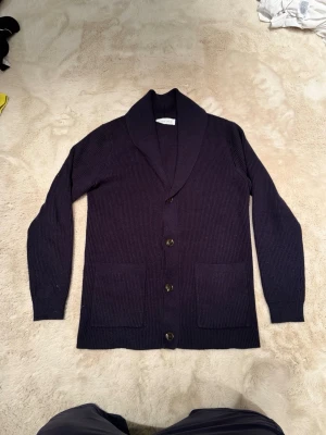 Mörkblå kofta från Pier One - Säljer en mörkblå ribbstickad kofta från Pier One. Koftan har sjalkrage, knäppning med fyra knappar framtill och två stora fickor. Perfekt för lager på lager under kyliga dagar.