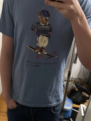 Blå Polo Bear t-shirt från Ralph Lauren - Snygg blå t-shirt från Polo Ralph Lauren med ikoniskt Polo Bear-tryck där björnen åker skidor. Modellen är custom slim fit och har korta ärmar. Tillverkad i mjuk bomull som känns skön mot huden. Perfekt för dig som gillar klassiska streetwear-vibbar.