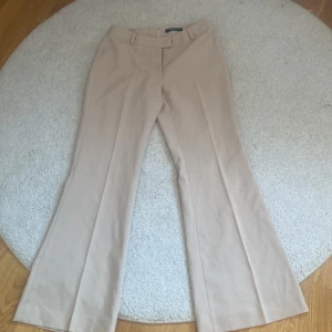 Beige kostymbyxor från Mandarin - Snygga beige kostymbyxor från Mandarini med diskret randigt mönster. Byxorna har mellan/hög midja. Perfekta för en clean och stilren look. Storleken är 36/38