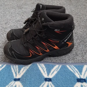 Svarta Salomon kängor med orange detaljer - Grymma svarta kängor från Salomon med orangea detaljer och robust sula. Skorna har snörning och är tillverkade i syntetmaterial med meshpaneler för extra komfort. Perfekta för utomhusaktiviteter och äventyr året runt.