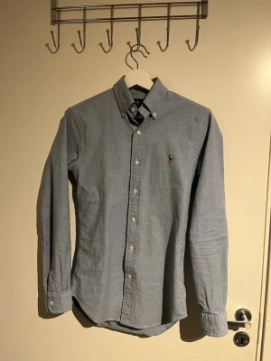 Blå slim fit skjorta från Ralph Lauren - Säljer en stilren blå skjorta från Ralph Lauren i slim fit-modell. Skjortan är i stretchig bomull, har klassisk button down-krage, långa ärmar och knappar framtill. Ikoniska Polo-loggan broderad på bröstet. Perfekt för dig som gillar en clean och tidlös look.