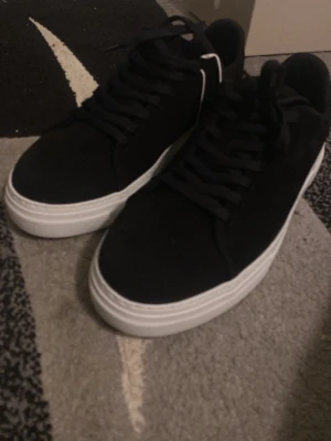 Svarta sneakers med vit sula - Stilrena svarta sneakers med tjock vit sula och klassisk snörning. Ovandelen är i mocka-look och designen är enkel och clean, perfekt till jeans eller shorts. Skorna har rund tå och platt sula för skön vardagsstil.