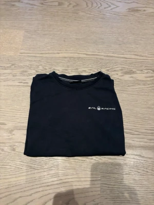 Svart t-shirt från Sail Racing - Svart t-shirt från Sail Racing med diskret logga på bröstet och en liten logga i nacken. Klassisk rund hals och korta ärmar. Tillverkad i mjuk bomull som känns skön mot huden. Perfekt för en clean och enkel look. FRAKT INGÅR EJ. Hör av dig ifall du vill se mer bilder. 