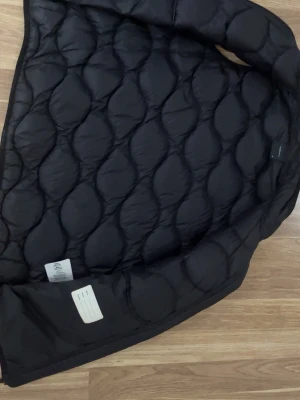 Svart dunväst Peak Performance - Säljer en svart dunväst från Peak Performance med quiltad design och hög krage. Västen har dragkedja framtill och två fickor med dragkedja. Perfekt för lager-på-lager och riktigt snygg till både hoodie och t-shirt.