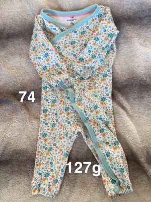 Blommig byxdress  - Supersöt blommig byxdress i vitt med turkosa, orange och rosa detaljer. Långärmad och helkroppsmodell med ljusblå kantband och knappar framtill. Perfekt för mysiga dagar hemma eller när du vill vara extra gullig.🌸 Tunnare variant. Fint använt skick. Storlek 74