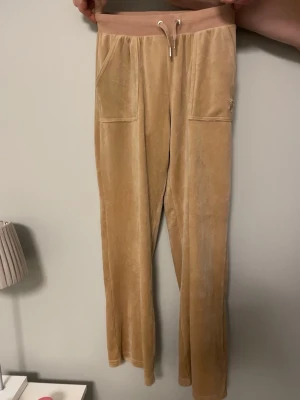 Jucy couture mjukis byxor barnmodell - Beiga mjukisbyxor från jucy couture i mjuk material med bred resårmidja och snörning. Byxorna har stora fickor fram och bakfickor med lock och knapp. Perfekta för chill dagar. Byxorna är 90 cm långa från midjan och 30 cm breda midja till midja dock så är byxorna elastiska så kan bli större samt 20 cm low waisted. Eftersom att byxorna är i barnmodell så finns det bara en knap på vardera ficka samt så är fickan lite mindre än vuxen storlek. Skriv till mig om du har några frågor🤗