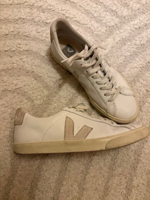 Vita Veja sneakers med beige detaljer - Säljer ett par klassiska Veja sneakers i vitt skinn med beige mockadetaljer på häl och sidor. Skorna har snörning och platt sula, samt den ikoniska V-loggan i mocka. Perfekta till jeans eller kjol för en clean och stilren look. Använd en sommar men inga stora defekter, storlek 38! Pris går att diskuteras!