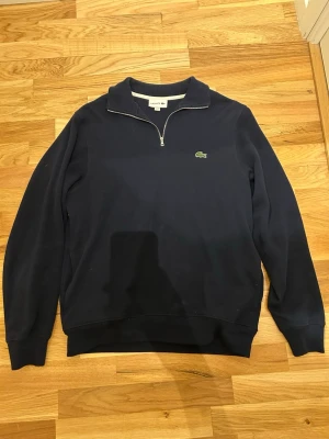 Mörkblå half zip tröja från Lacoste - Snygg mörkblå tröja från Lacoste med halv dragkedja och klassisk krage. Tröjan har lång ärm och den ikoniska gröna krokodilloggan på bröstet. Perfekt för dig som gillar en stilren look.