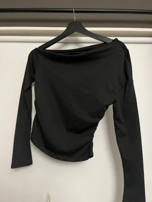 Svart långärmad topp med bred hals - En stilren svart långärmad topp med bred, som bara täcker en axel. Toppen har en figurnära passform och är tillverkad i ett mjukt, stretchigt material som ger en clean look. Perfekt att matcha med jeans eller kjol för en enkel men snygg outfit.