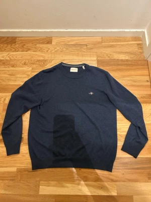 Mörkblå stickad tröja från GANT - Säljer en mörkblå stickad tröja från GANT i storlek M. Tröjan har rund hals med diskreta ränder i kragen och GANT-logga broderad på bröstet. Klassisk passform och långa ärmar, perfekt för lager på lager under höst och vinter.