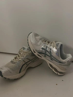 Vita och silver Asics sneakers - Säljer ett par riktigt snygga Asics sneakers i vitt och silver med mesh och syntetmaterial. Skorna har klassisk snörning, coola detaljer och en rejäl sula med bra grepp. Perfekta för dig som gillar sportig stil och vill ha bekväma skor till vardagen. Har ej kvitto för har fått dom! 