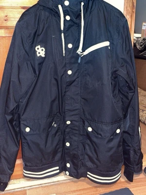 Svart vindjacka från Dope, strl M - Svart vindjacka från Dope med vita detaljer och dragkedjor. Jackan har huva med snörning, flera fickor med knappar och dragkedja, samt ribbade muddar med vita ränder. Insidan har mesh och logoband. Perfekt för dig som gillar streetwear och funktion. Samt ventilation vid båda sidor vid armen, sen liftkort ficka vid sidan av armen. En knapp saknas på framsidan.