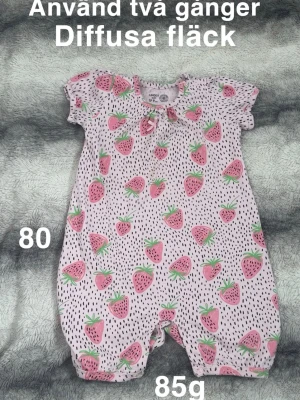 Rosa byxdress med jordgubbsmönster - Supersöt rosa byxdress från Lindex med jordgubbsmönster och svarta prickar. Kortärmad modell med knappar i grenen för enkel påklädning. Perfekt för varma dagar och lek!🌸 Använd två gånger men fått diffusa fläckar. Storlek 80