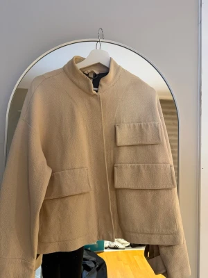 Beige overshirt med fickor XS - Säljer en beige overshirt i storlek XS med coola stora fickor framtill och ståkrage. Jackan har en loose passform och är tillverkad i ett strukturerat material som känns mjukt och skönt. Perfekt att slänga på över en hoodie eller t-shirt.