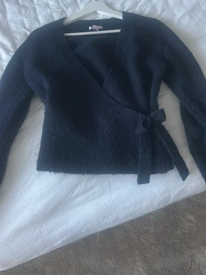 mörkblå omlottkofta från Gina Tricot - Mysig svart stickad omlottkofta från Gina Tricot med knyt i sidan. Koftan har lång ärm och en fluffig känsla, perfekt för kyliga dagar. Enkel att styla med jeans eller kjol för en chill look. köpt för 400 därav priset 