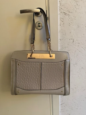 Grå handväska från River Island - Snygg ”greige” handväska från River Island med guldfärgade detaljer och kedjor. Knappast använd. Väskan har präglat mönster med RI-logga framtill och stilrena svarta kanter. Perfekt storlek, klassisk och trendig design.