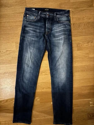 Mörkblå jeans från Jack & Jones - Säljer ett par mörkblå jeans från Jack & Jones med snygg tvättad effekt och klassisk femficksdesign. Jeansen har normal passform och raka ben, perfekta till en avslappnad stil. Materialet är slitstarkt jeans och färgen har en schysst ljusare tvätt framtill. 28/32