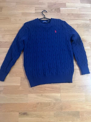 Ralph Lauren tröja  - Säljer en marinblå kabelstickad tröja från Polo Ralph Lauren i storlek M. Tröjan har rund hals, långa ärmar och den klassiska röda broderade loggan på bröstet. Perfekt för kyliga dagar och enkel att matcha med jeans eller chinos.