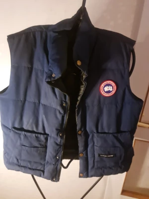 Mörkblå dunväst från Canada Goose - Snygg mörkblå dunväst från Canada Goose med klassisk logga på bröstet. Västen har två stora fickor framtill, dragkedja och tryckknappar. Perfekt för lager-på-lager och håller dig varm under kyliga dagar.
