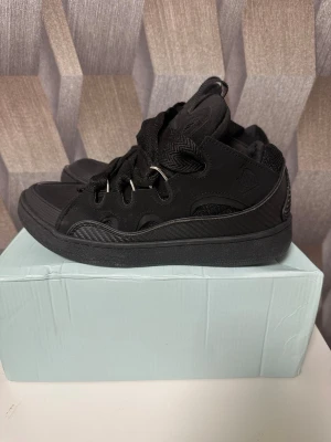 Svarta lanvins curb - Säljer ett par helsvarta sneakers med chunky design och breda, mönstrade snören. Skorna har detaljer i mocka och mesh, samt metallhål för snörningen. Snyggt sportig look med rund tå och platt sula. Perfekta för dig som gillar streetwear.använda bara ett fåtal gånger annars jätte bra skick