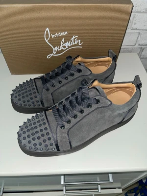 Gråa Christian Louboutin sneakers med nitar - Säljer ett par gråa sneakers från Christian Louboutin i mocka med svarta nitar på tån och svarta snören. Skorna har den ikoniska röda sulan och coola detaljer som gör dem till ett statement. Perfekta för dig som vill sticka ut med lyxig streetstyle.