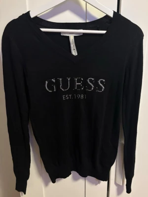 Svart långärmad tröja från Guess - Svart långärmad tröja från Guess med v-ringning och glittrande stenar som bildar loggan på bröstet. Tröjan är i tunn, mjuk kvalitet och har en klassisk passform. 