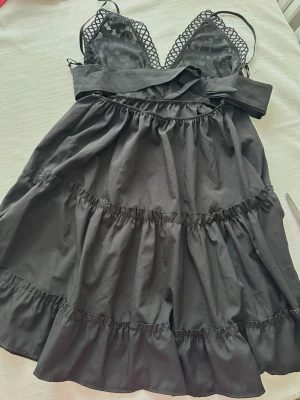 Svart spetsklänning från Koyye M  - Supersöt svart kortklänning från Koyye med spetsdetaljer upptill, smala axelband och omlottknytning i midjan. Kjolen är volangig och har flera lager för extra fluff. Perfekt för dig som gillar en feminin och trendig look.