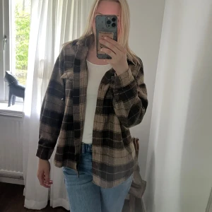 Brun flanell skjorta overshirt - Säljer en brun och svart skjorta med klassisk krage och knappar framtill. Skjortan har lång ärm och är tillverkad i ett mjukt material som känns skönt mot huden. Perfekt att slänga över en t-shirt för en chill look.