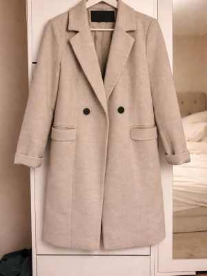 Kappa - Snygg beige kappa från Vero Moda med klassisk krage och dubbelknäppning framtill. Jackan har två fickor med lock och är helfodrad. Perfekt för dig som vill ha en stilren och tidlös look under kyligare dagar.