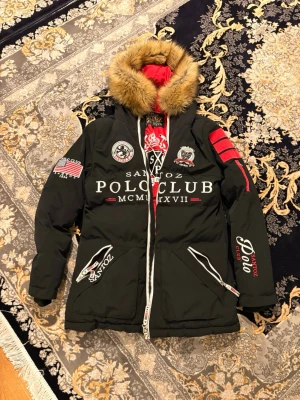 Svart pufferjacka från Santoz Polo Club - Svart pufferjacka från Santoz Polo Club med broderade detaljer, stor huva med päls och röda inslag på insidan. Jackan har dragkedja framtill, två snedställda fickor med dragkedja och flera broderade loggor och märken på bröstet och ärmarna. Pris kan diskuteras vid snabb affär.