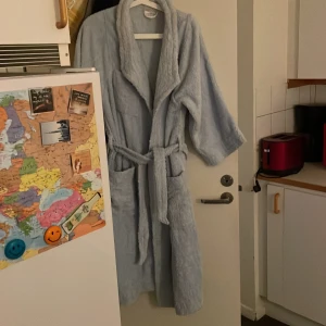 Ljusblå badrock i frotté från Penella - Mysig ljusblå badrock i frotté från Penella. Modellen har sjalkrage, stora fickor och knytskärp i midjan. Perfekt att slänga på sig efter duschen eller när du chillar hemma. Broderad detalj på bröstet för en extra touch.Lite tyngre morgonrock med 3/4 ärmar.Använd sparsamt till gäster.