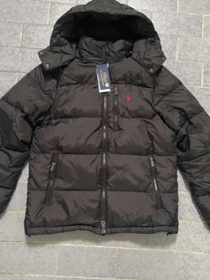  Polo Ralph Lauren jacka - Svart pufferjacka från Polo Ralph Lauren med huva och flera dragkedjor framtill. Jackan har ett rött broderat Polo-logo på bröstet och är tillverkad i polyester. Perfekt för kalla dagar och har en snygg, sportig vibe.