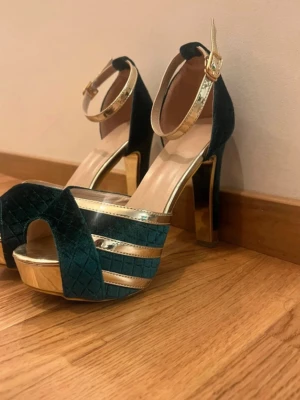 Gröna platåklackar med guldiga detaljer - Säljer ett par gröna platåklackar med quiltad sammet och guldiga metallicdetaljer. Skorna har öppen tå, hög klack och justerbart ankelband i guld. Perfekta för dig som vill sticka ut med både färg och höjd. Snyggt rutmönster på ovandelen och stabil plattform.