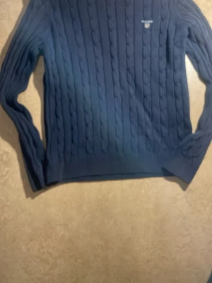 Mörkblå kabelstickad tröja från GANT - Säljer en mörkblå kabelstickad tröja från GANT med rund halsringning och diskret logga broderad på bröstet. Tröjan har långa ärmar och ribbade muddar vid ärmslut och nederkant. Perfekt för dig som gillar klassisk och stilren look.