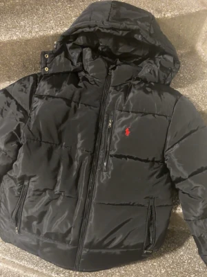Svart pufferjacka från Polo Ralph Lauren - Säljer en svart pufferjacka från Polo Ralph Lauren med avtagbar huva och klassisk röd logga på bröstet. Jackan har flera dragkedjeförsedda fickor och justerbara band i midjan. Perfekt för kalla dagar och riktigt snygg till streetwear.
