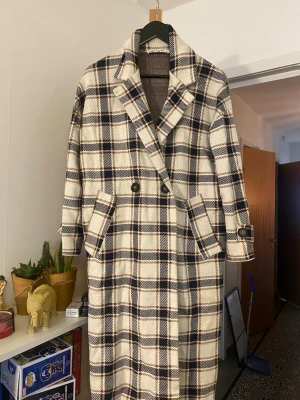 Rutig lång kappa från Zara - Säljer en snygg lång kappa från Zara i beige, svart och brunt rutigt mönster. Kappan har dubbelknäppning, bred krage och fickor med lock. Perfekt för dig som vill ha en statement jacka till höst och vinter.