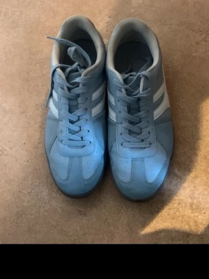 Blå sneakers med vita detaljer - Snygga blå sneakers med vita ränder på sidorna och matchande blå snörning. Skorna har en platt svart sula och är tillverkade i syntetmaterial med en sportig vibe. Perfekta för dig som gillar en clean och stilren look.