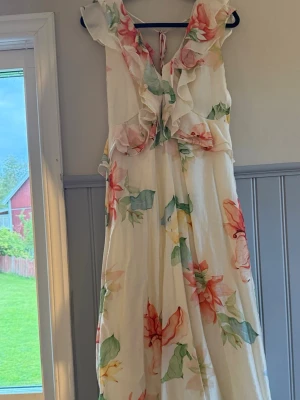 Blommig långklänning med volang - Superfin långklänning i ljus bas med stora blommor i grönt, rött och orange. Klänningen har volangdetaljer vid axlar och byst, v-ringning och knyt i ryggen. Perfekt för sommarens alla tillfällen. Materialet känns lätt och luftigt. Använd fåtal gånger❤️‍🔥
