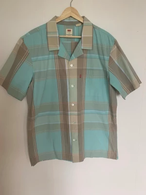 Rutig kortärmad skjorta från Levi's - Snygg rutig skjorta från Levi's i ljusblått och beige med klassisk krage och korta ärmar. Skjortan har knappar framtill och en liten bröstficka med Levi's-röd etikett. Perfekt för chill dagar och sommarhäng.