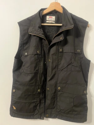 Svart väst från Fjällräven - Säljer en svart väst från Fjällräven med flera praktiska fickor och tryckknappar. Västen har hög krage, dragkedja och knappstängning framtill. Perfekt för dig som gillar funktionella plagg med snygg design.