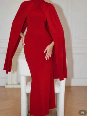 Röd cape-klänning med lång kjol - Snygg röd långklänning med cape-detalj som ger en dramatisk look. Klänningen har figurnära passform och hög hals, samt långa, svepande ärmar som skapar en elegant siluett. Perfekt för dig som vill sticka ut på nästa event.