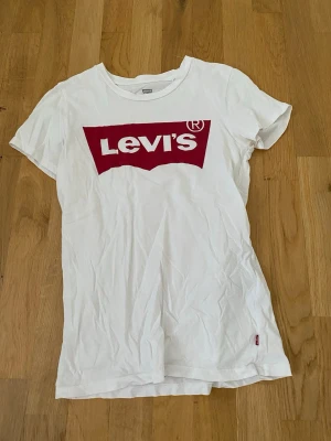 Vit Levi's t-shirt med röd logga XS - Klassisk vit t-shirt från Levi's i storlek XS med den ikoniska röda loggan tryckt på bröstet. T-shirten har rund hals, korta ärmar och är gjord i mjuk bomull. Perfekt till jeans eller shorts för en enkel och snygg look.