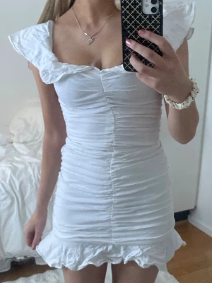 Vit kortklänning/studentklänning med volang - Supersöt vit kortklänning med volangkant nertill och breda axelband. Klänningen har rynkade detaljer längs hela kroppen och en feminin, figurnära passform. Perfekt för sommaren eller studenten. Ganska petit i storlek, skulle säga xs.