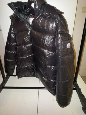 Svart Moncler pufferjacka  - Svart pufferjacka från Moncler med glansig finish och klassisk logga på ärmen. Jackan har huva, dragkedja framtill och praktiska fickor. Insidan har en cool serietidningsetikett. Perfekt för dig som vill ha en varm och snygg jacka med streetkänsla. !!Denna annons includerar en valfri moncler t-shirt!!