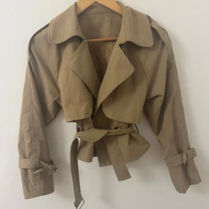 Beige croppad trenchcoat med bälte - Snygg croppad trenchcoat i beige med bred krage, dubbelknäppning och bälte i midjan. Jackan har långa ärmar med spännen vid handlederna och klassiska trench-detaljer. Perfekt för dig som vill ha en trendig och stilren look.